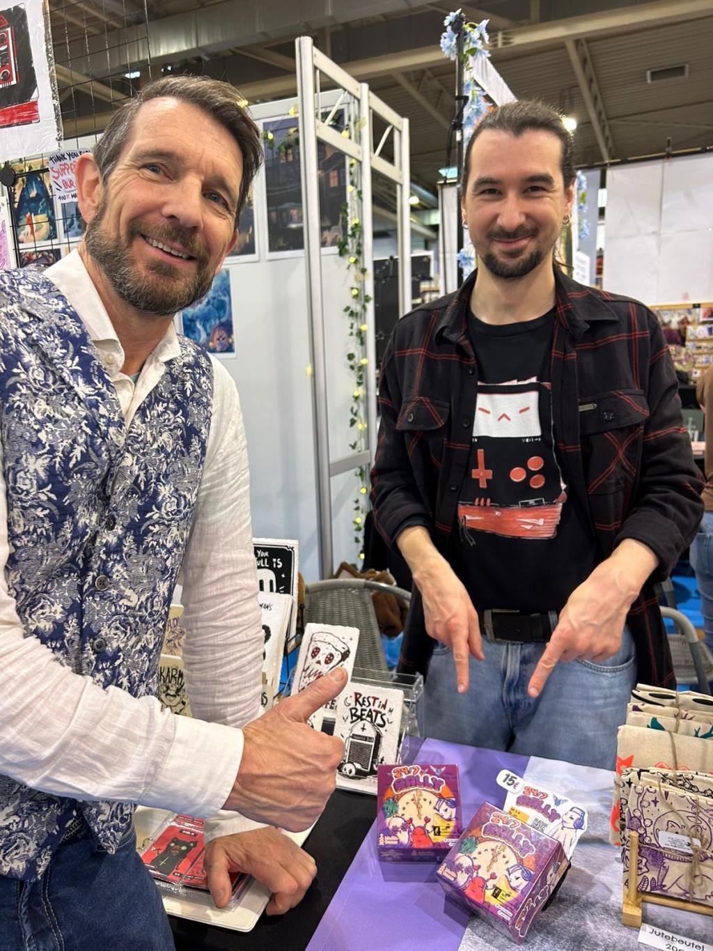 Jan Stronkman en Thiemo Müller wijzen lachend naar het spel 24/7 Rally tijdens de Spiel Essen beurs.