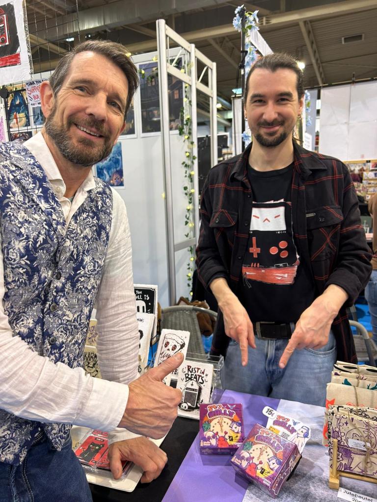 Thiemo Müller van Tintenkrach samen met Jan Stronkman van Madilo.games wijzen naar het familiespel 24/7 Rally tijdens de beurs Spiel25 in Essen.